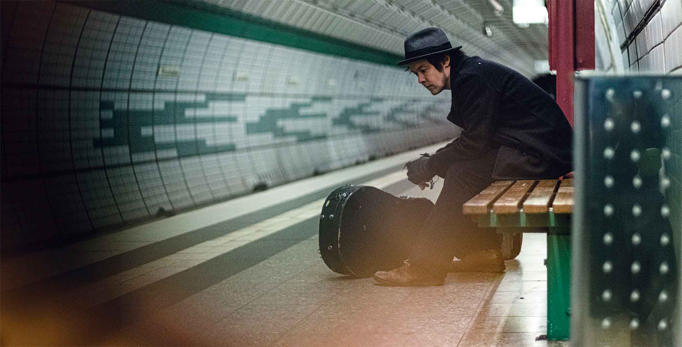 picture of jon sitting in the subway station on a bench with the guitar bag in front of him landscape mode